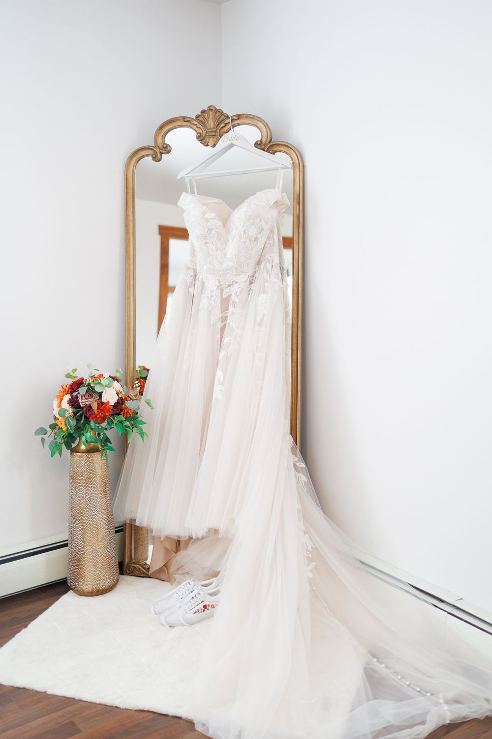 A delicate white wedding dress with flowing tulle and lace details hangs elegantly against ornate gold-framed mirrors.