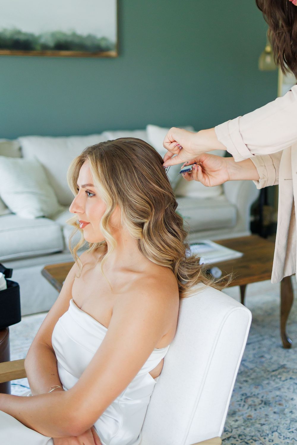 Stylist arranges a bride's hair while she sits in a white chair during wedding preparations.