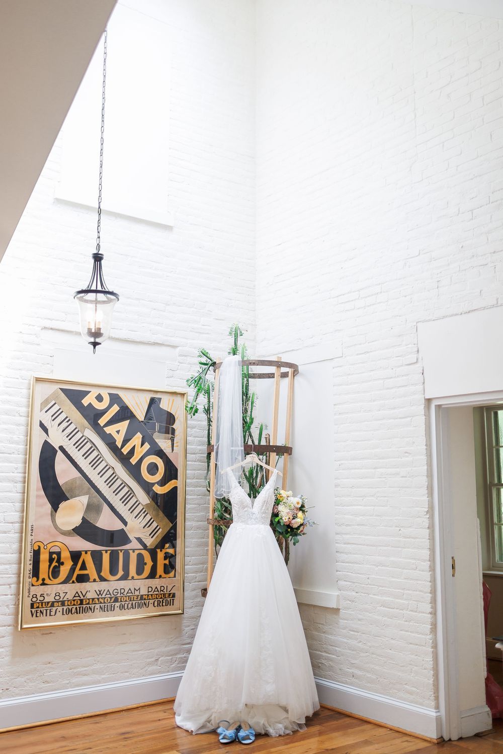 A wedding dress hangs elegantly in a white brick room with vintage art deco style posters on the wall.