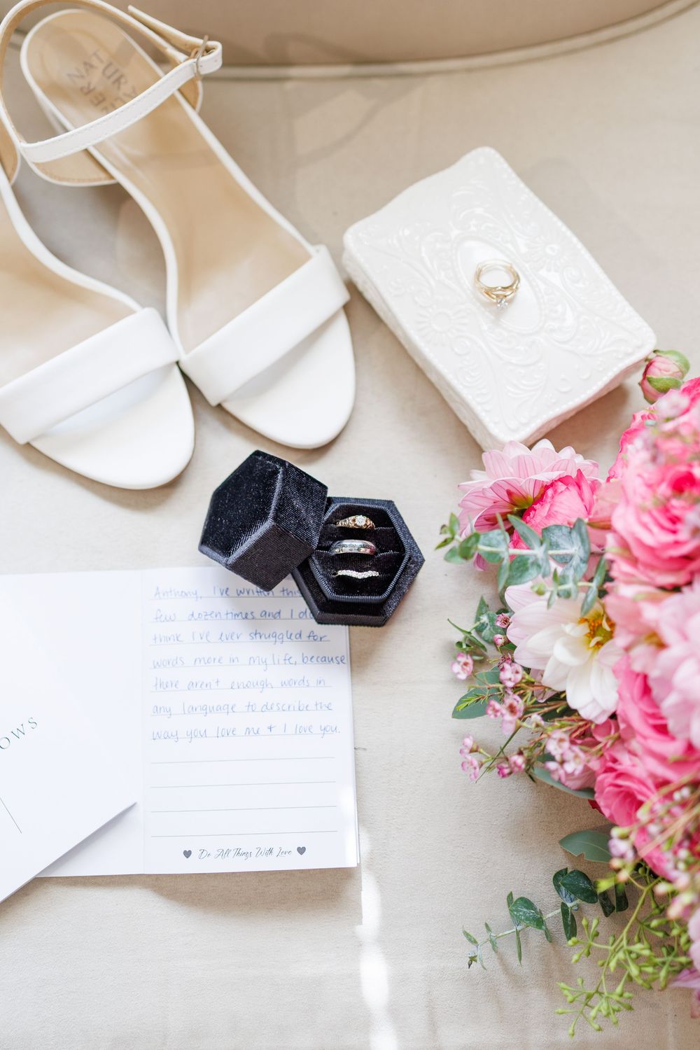 Wedding details flatlay with pink roses, white shoes, rings in black hexagon box and handwritten notes on beige background.