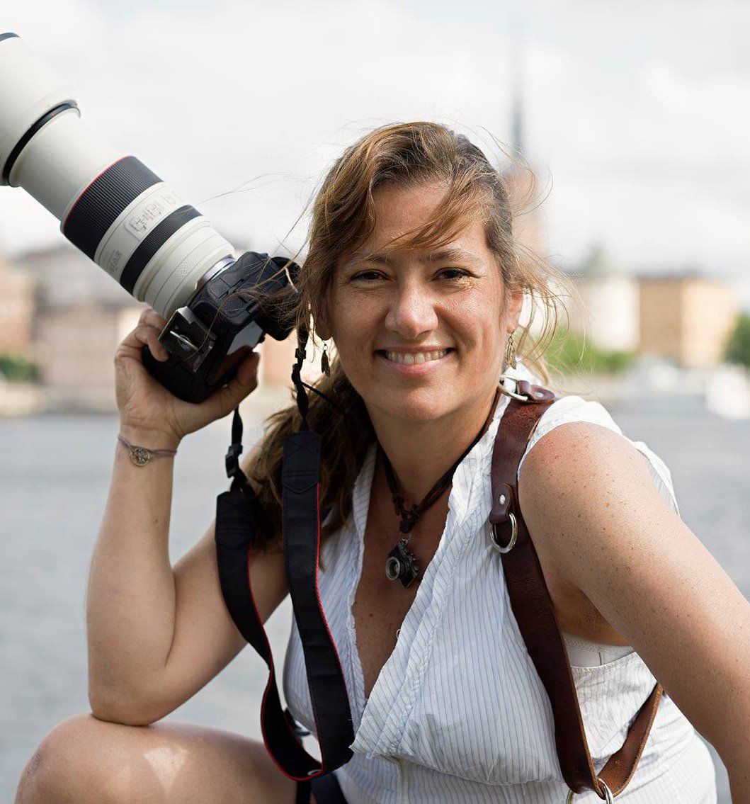 A photographer with a professional camera and telephoto lens smiles while taking outdoor photos on a sunny day.