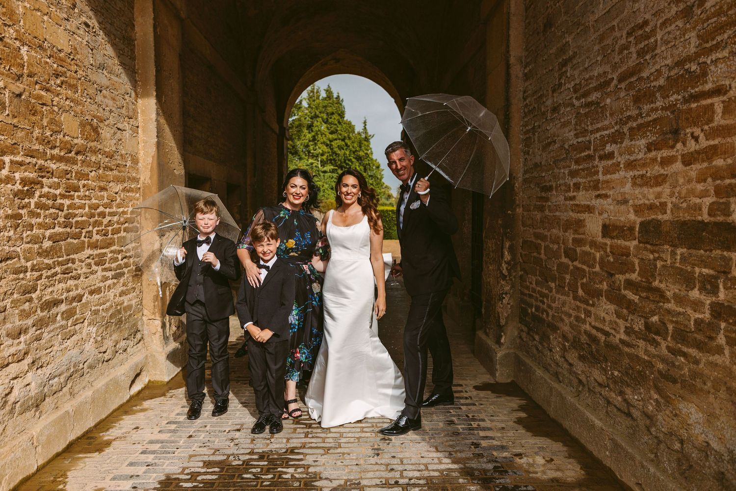Wedding party walks together under a stone archway while holding umbrellas on a rainy day.