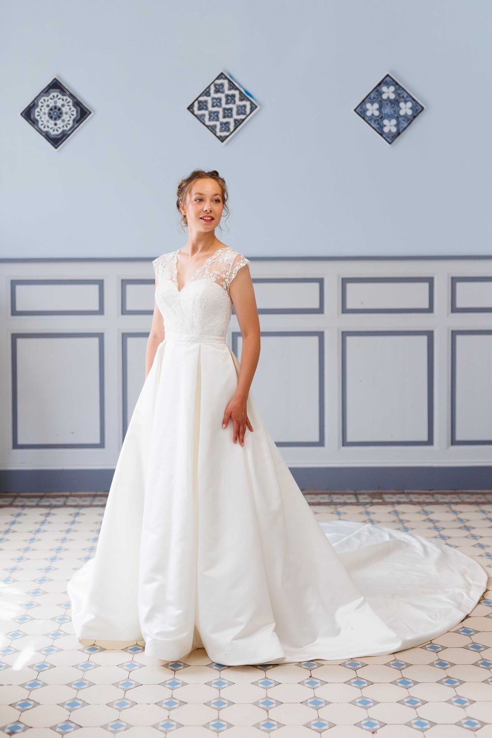 A wedding dress with a flowing train displayed on a tiled floor with decorative wall art in the background.