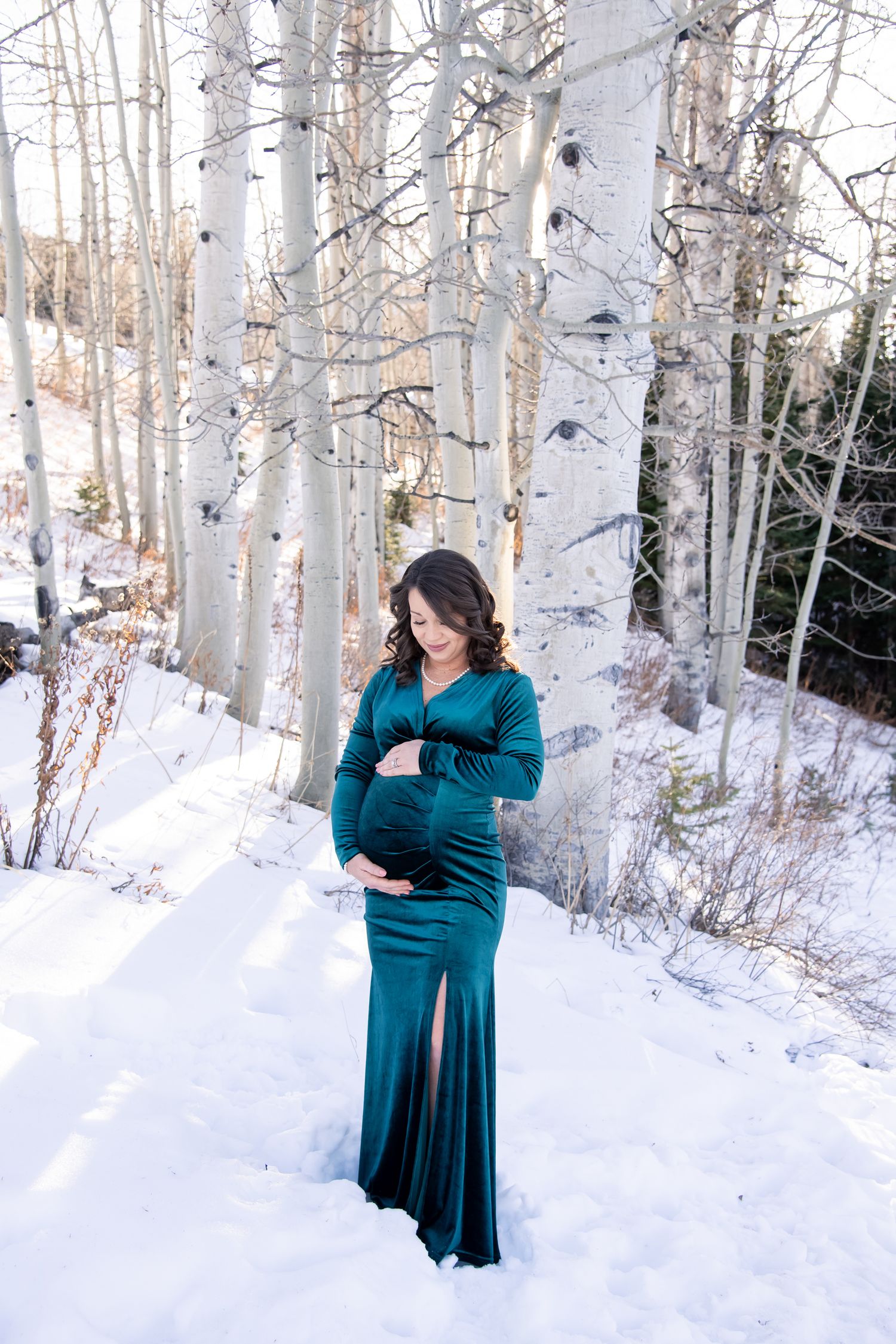 A woman in a teal dress poses gracefully among snow-covered birch trees in a winter forest setting for maternity photo session in Aspen CO