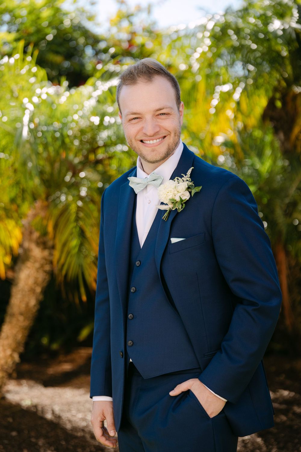Groom in navy suit and white bowtie poses in a lush tropical garden setting.