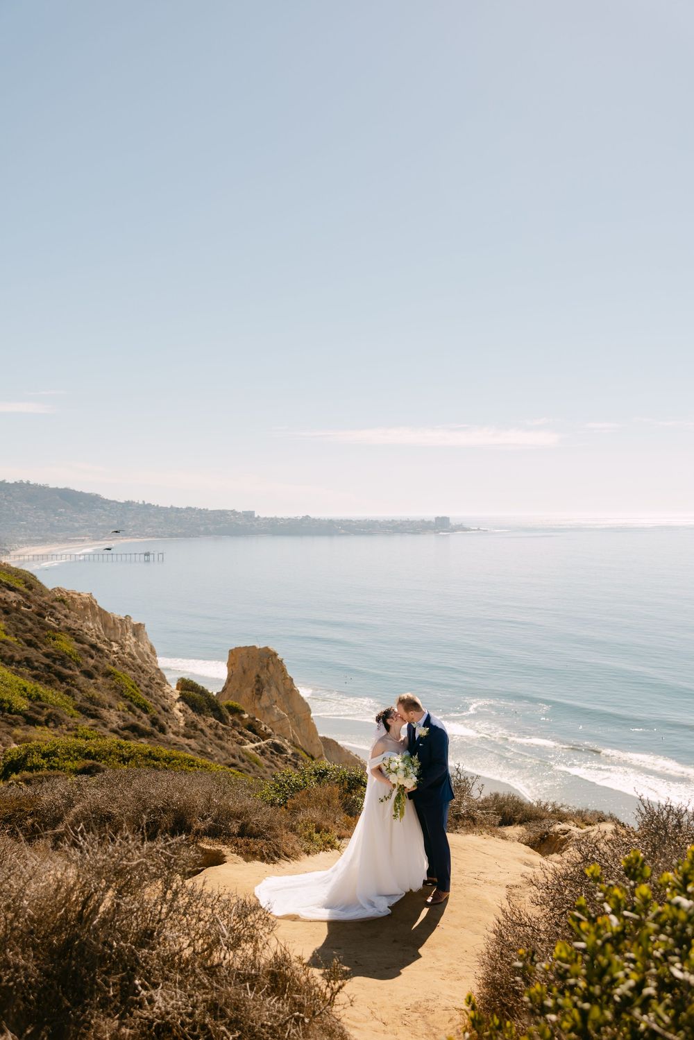 A couple shares an intimate moment on a coastal cliff overlooking the Pacific Ocean on a sunny day.