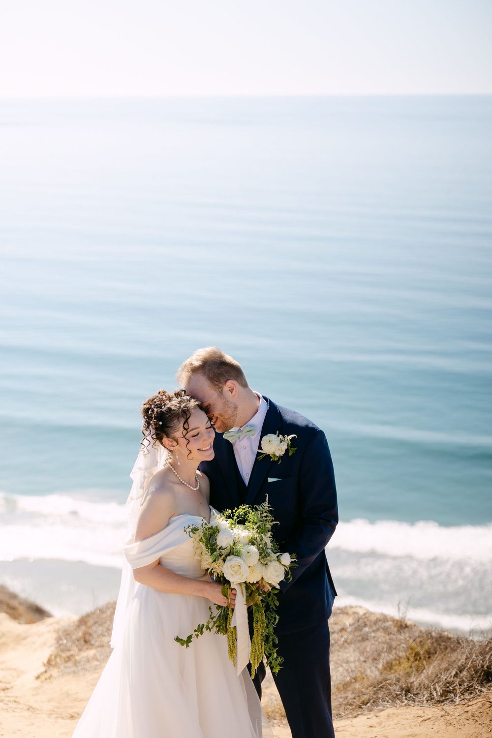A romantic coastal wedding photo shoot with a couple embracing on a cliff overlooking the ocean.