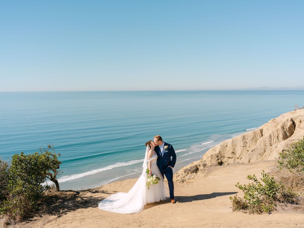 Wedding couple poses on coastal cliffs overlooking a pristine blue ocean.