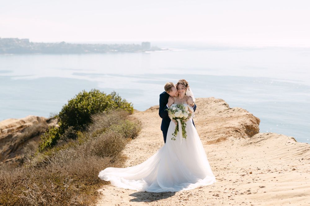 A couple in wedding attire stands on a scenic coastal cliff overlooking the ocean on their special day.