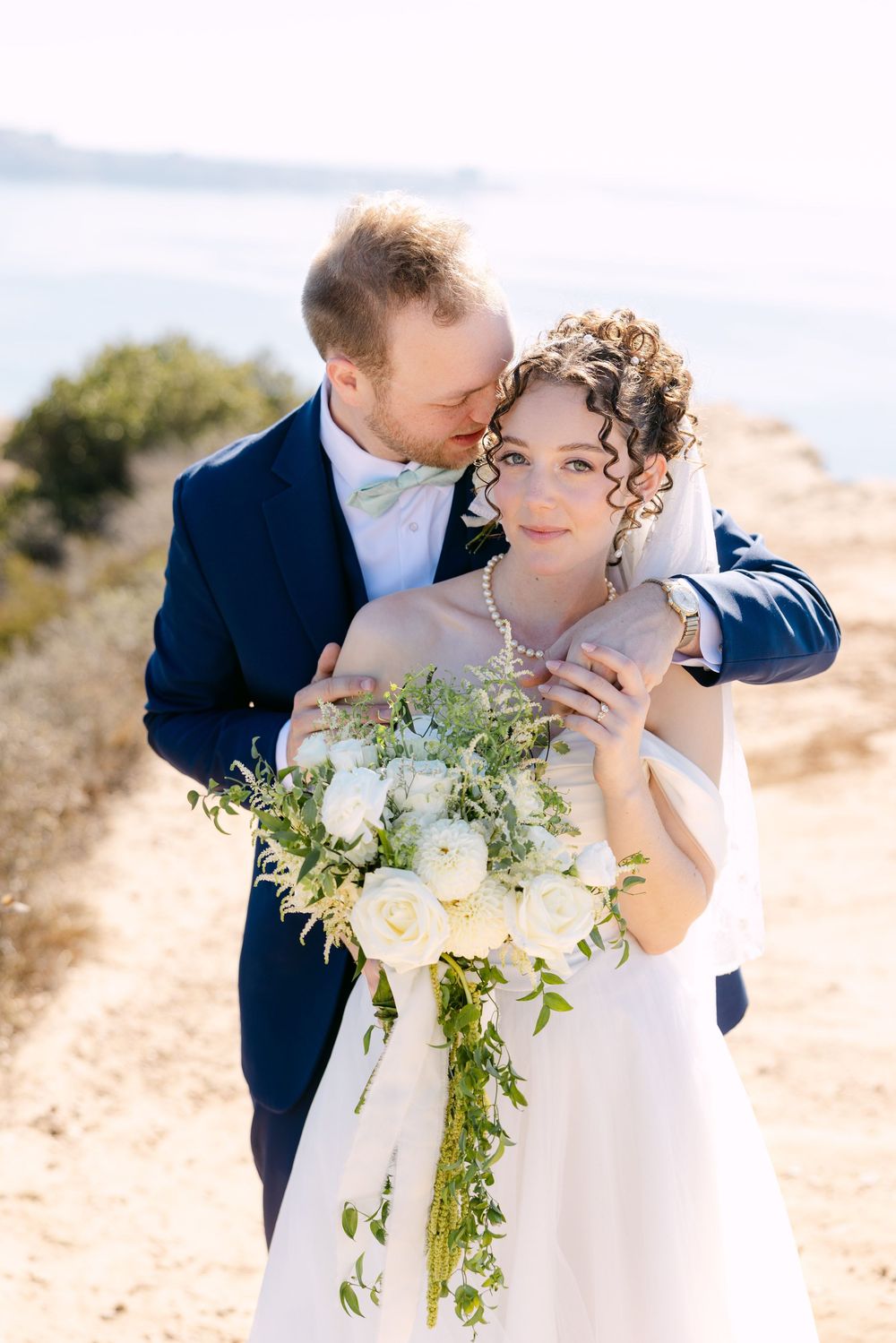 A wedding couple shares intimate moments on a sunny coastal cliff with white floral bouquet.