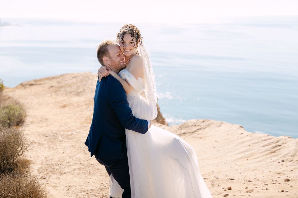 A couple embraces on a scenic coastal cliff overlooking the ocean during their wedding photoshoot.