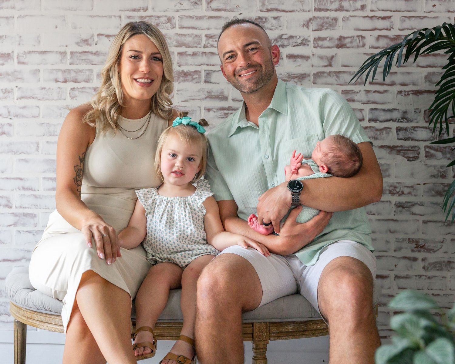 Happy family of four posing for newborn portrait, parents with toddler girl and newborn baby against white brick wall.