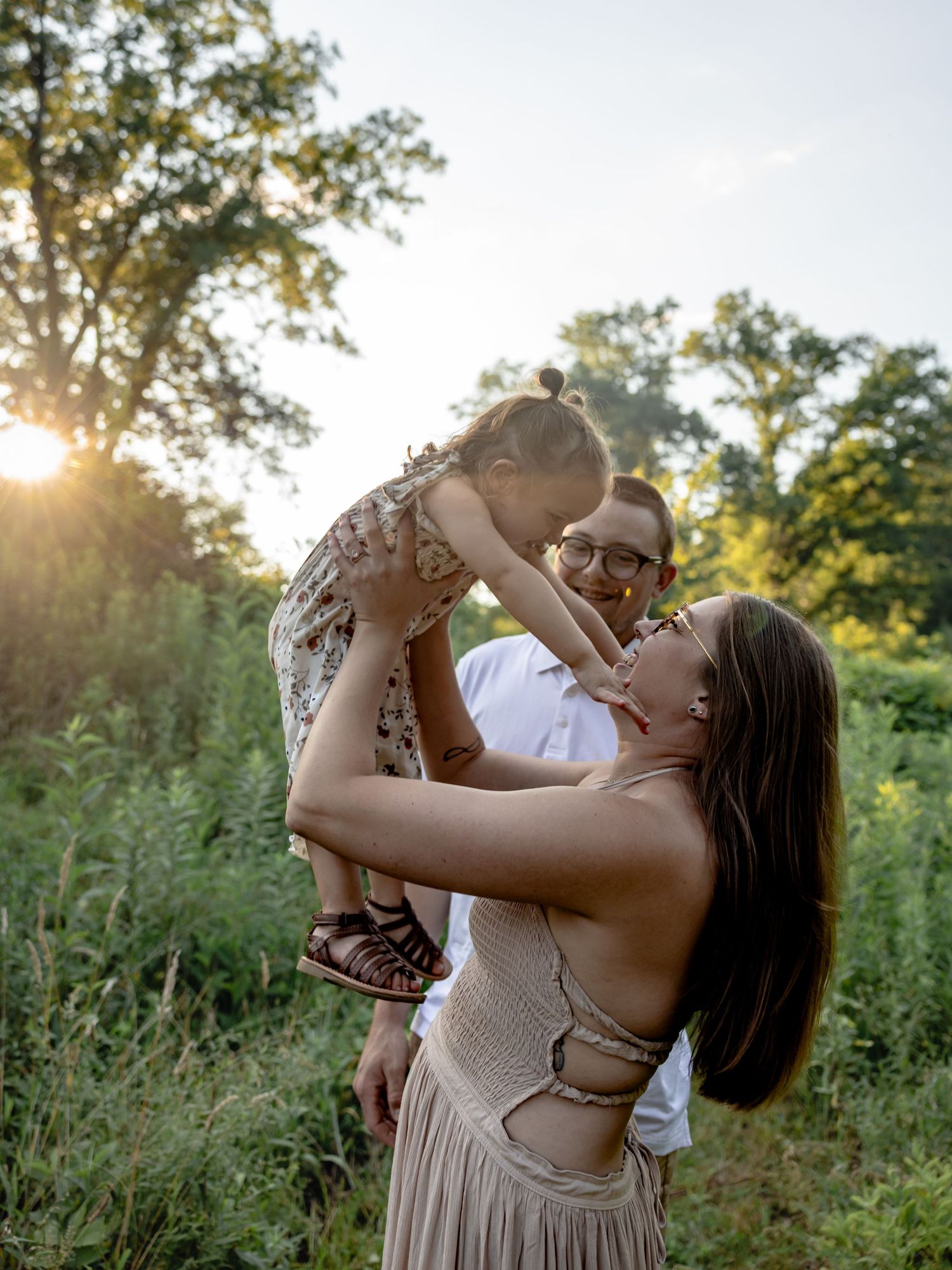 A couple lifts their toddler in a sunlit forest clearing, sharing a joyful family moment at golden hour.