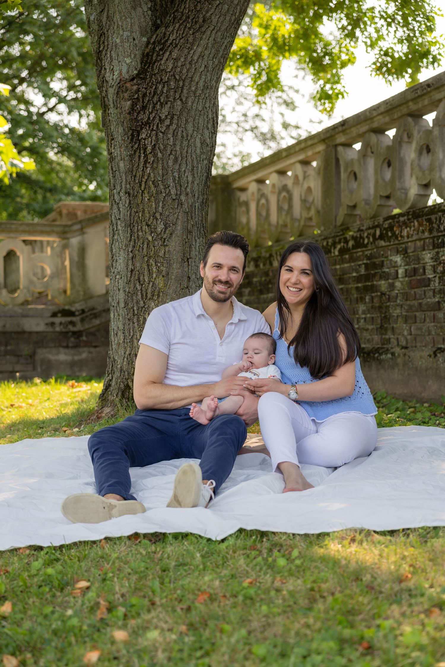 A happy young family with a baby sits on a white blanket under a large tree in a lush green park.