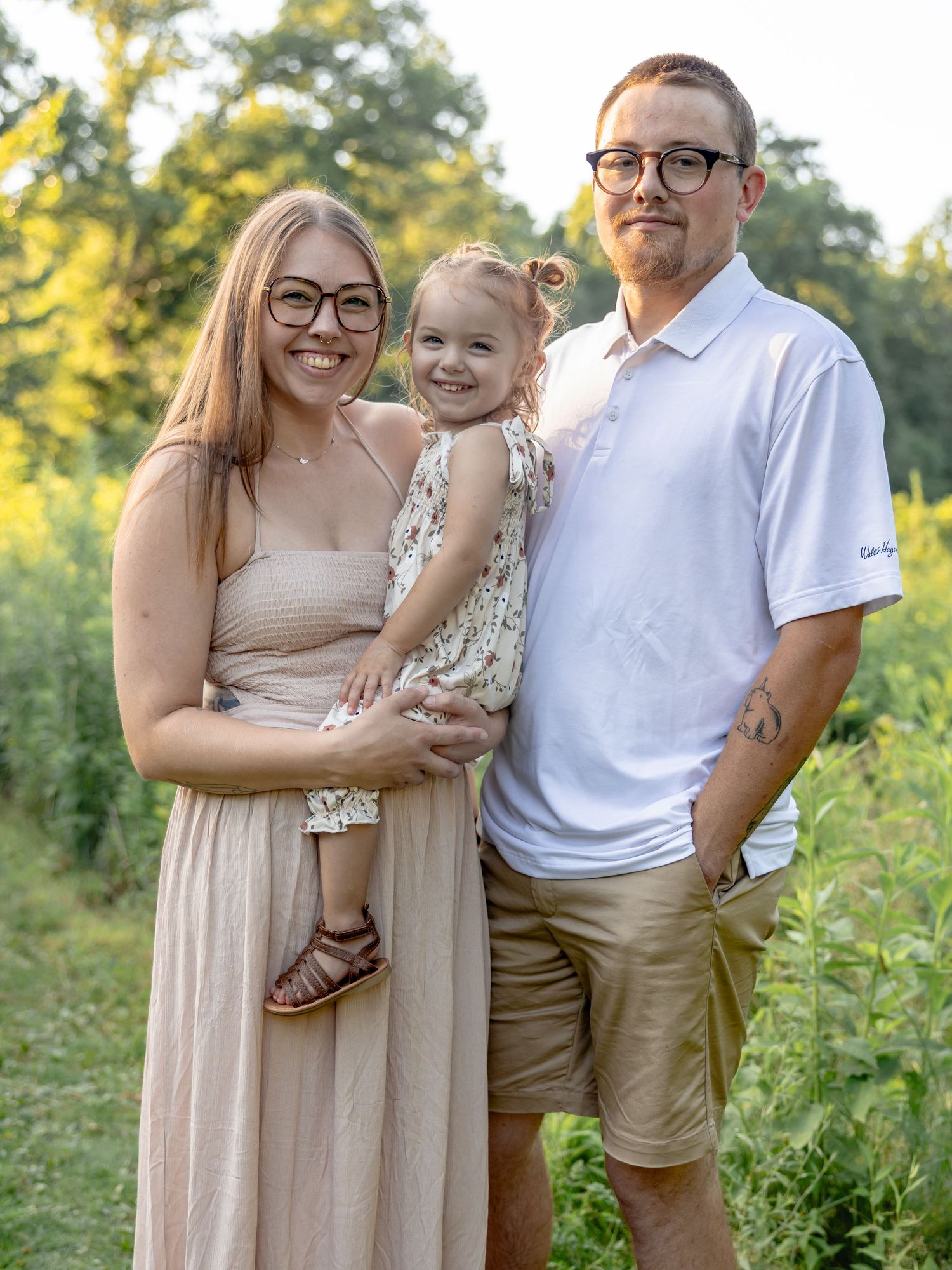 A smiling family of three poses outdoors in a sunny green field, with parents holding their young daughter between them.