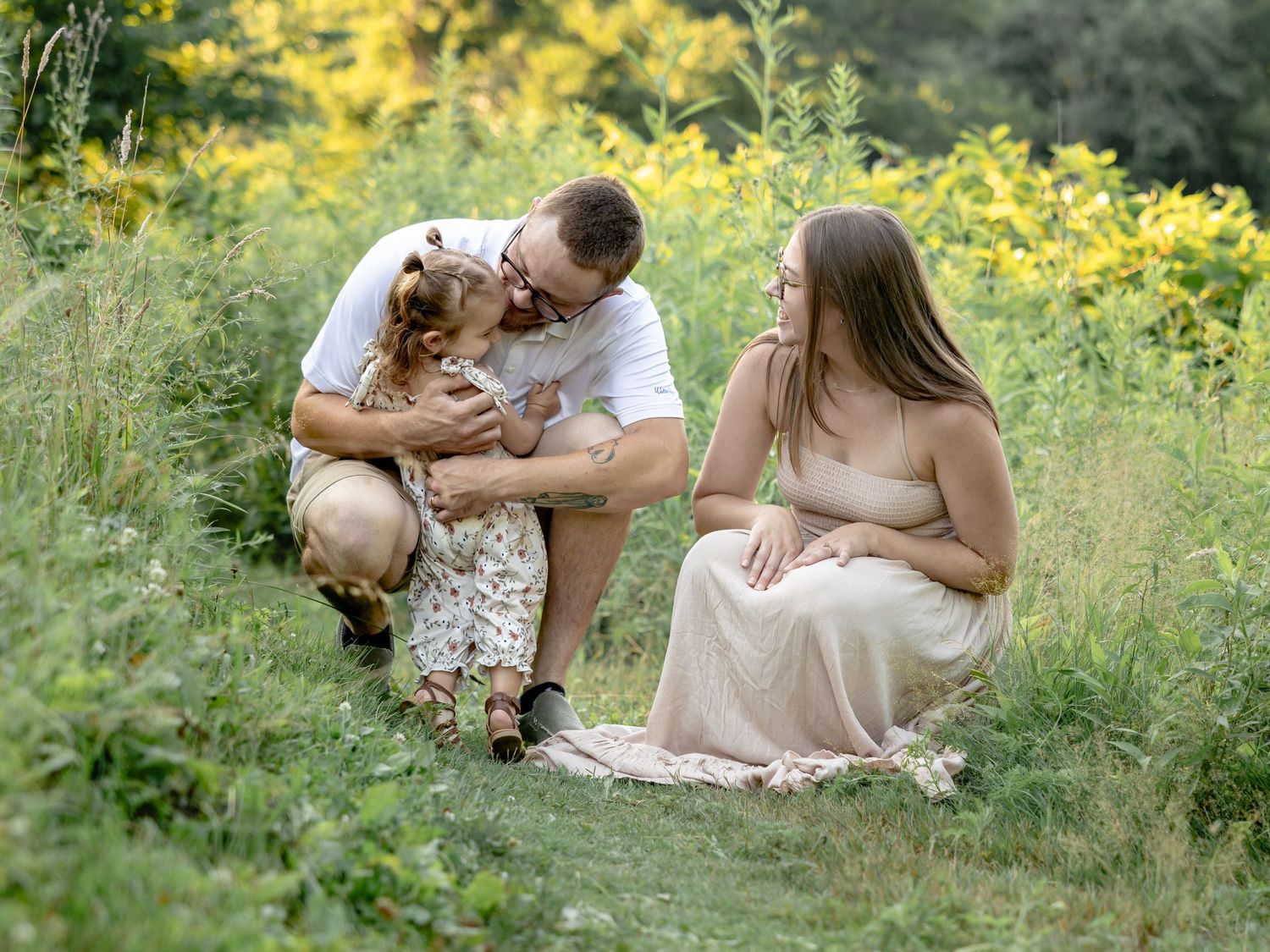 Pregnant woman in blush gown sits outdoors with husband and toddler daughter among wildflowers in a maternity photo.