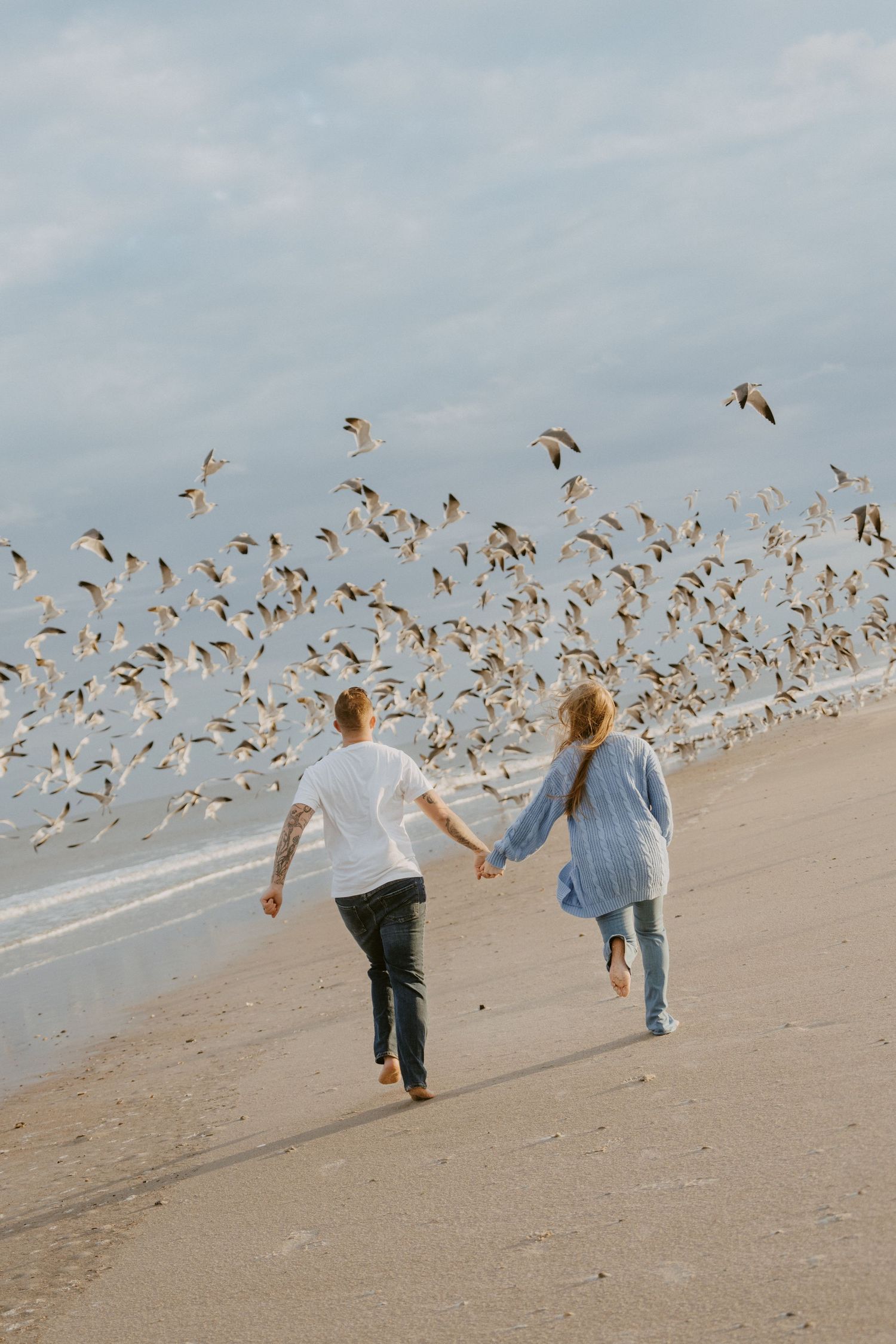 A couple in white attire walks along a sandy beach path at sunset with palm trees in the background.
