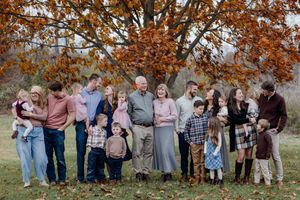 Large group of people in casual attire gathered outdoors for family photos during autumn season with orange leaves.
