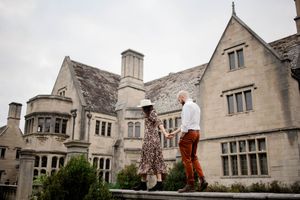 Couple holding hands outside a grand stone manor house, woman in floral dress and man in rust trousers.