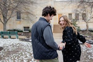 A young couple laughs and holds hands outdoors on a snowy winter day near a stone building.