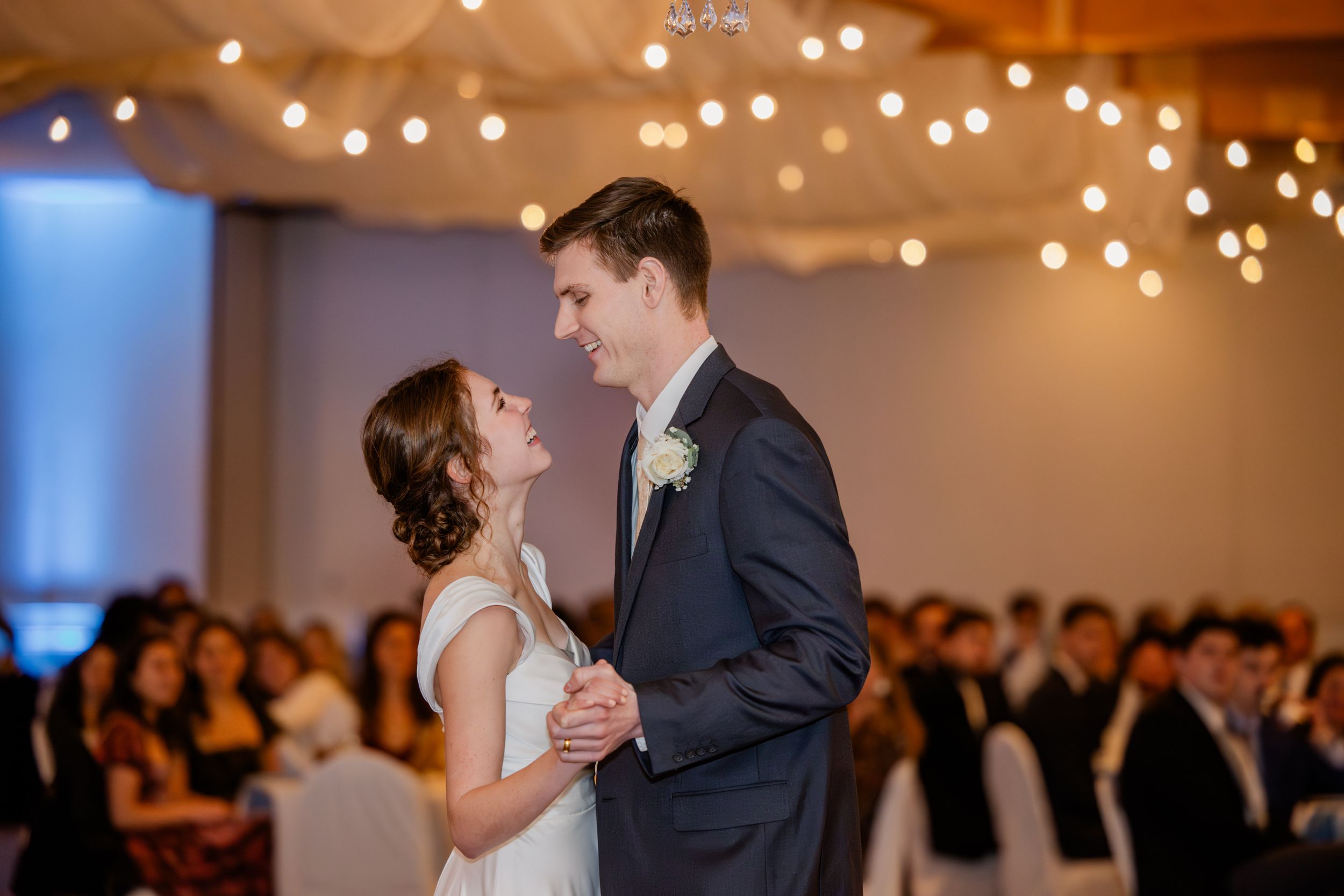A couple shares their first dance at a wedding reception under warm glowing lights and surrounded by guests.