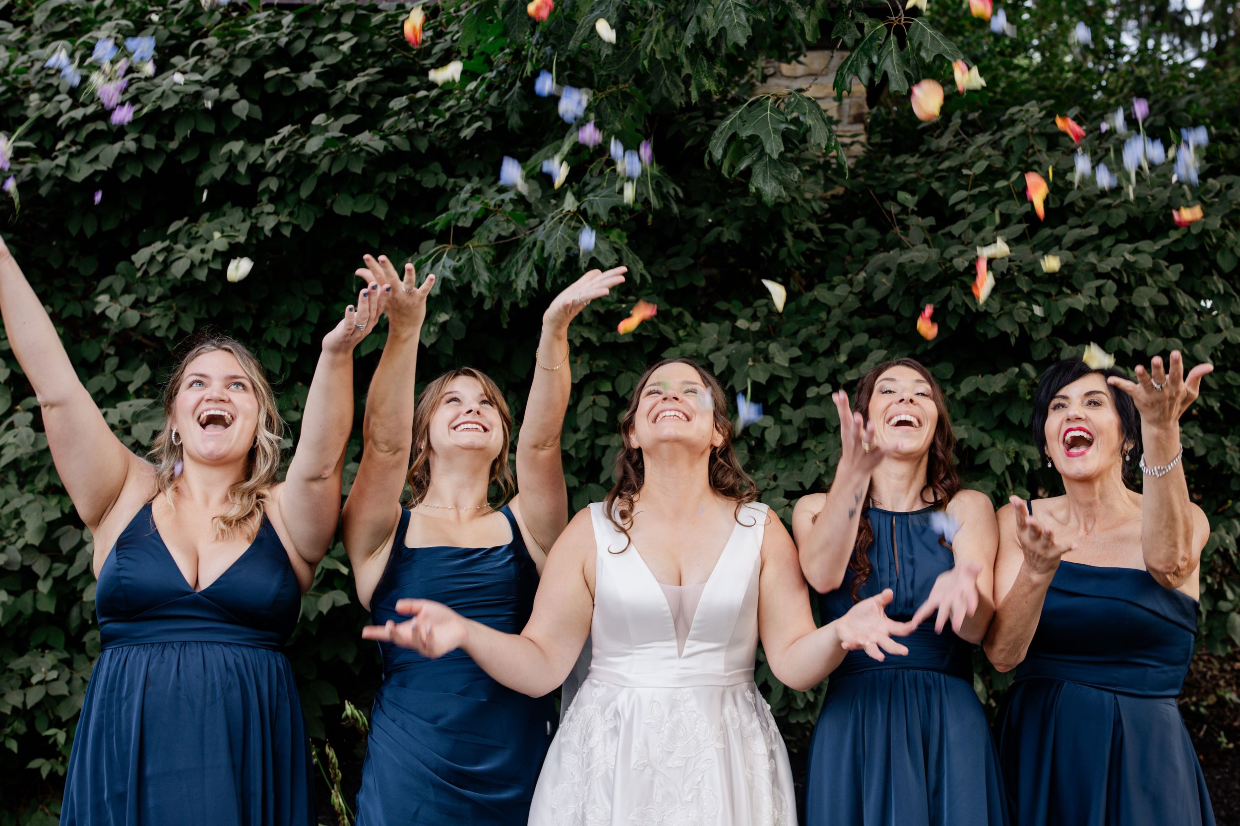 Bride in white gown and bridesmaids in navy dresses laughing as colorful flower petals are tossed in the air outdoors.