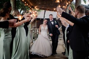 Bride and groom walk through sparkler send-off with wedding party in sage green dresses and black tuxedos.