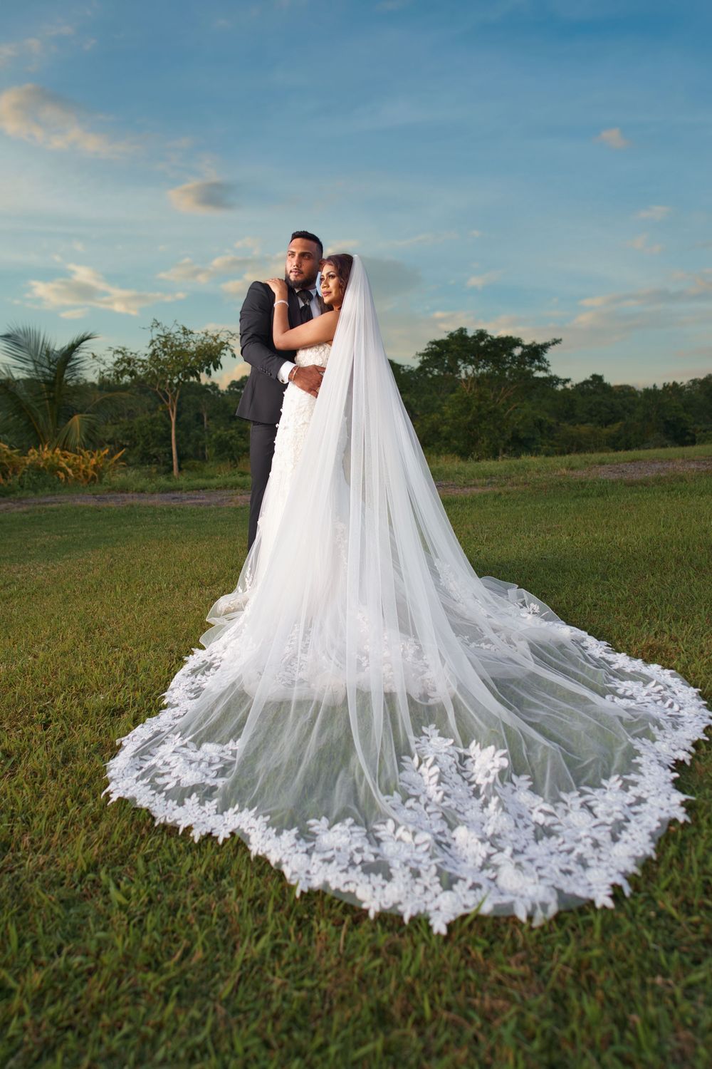 A wedding couple embraces in a grassy field at sunset with the bride's long lace-trimmed cathedral veil flowing behind them.