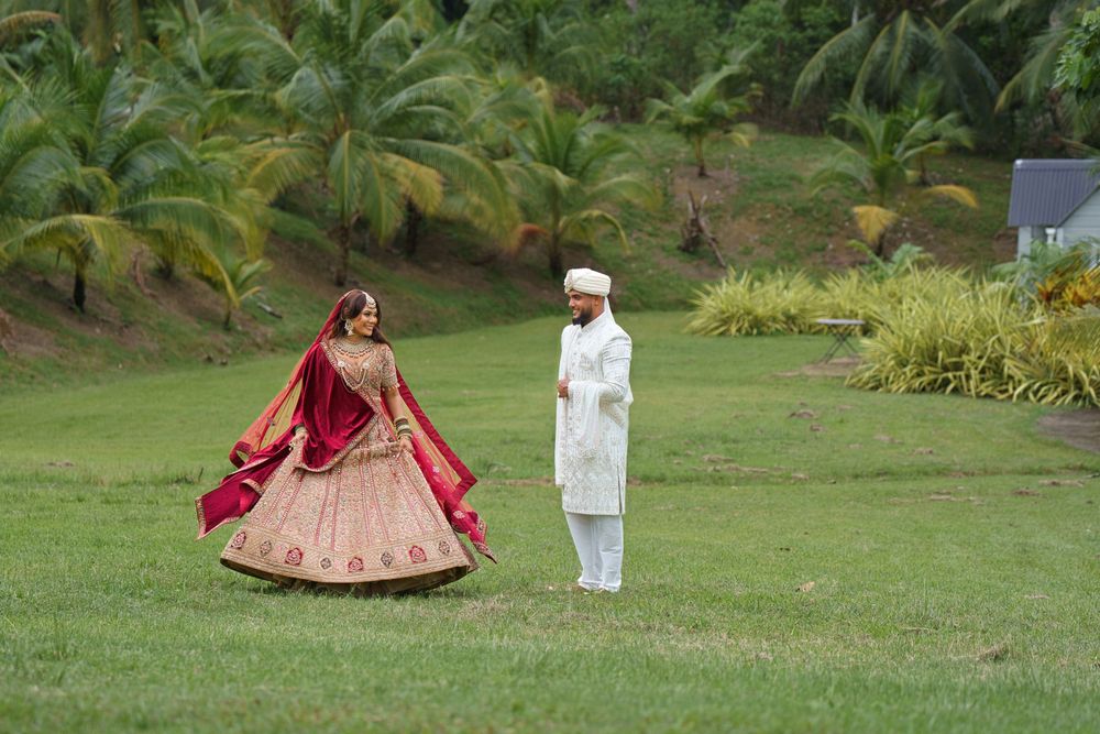 A couple poses in traditional Indian wedding attire on a lush green lawn with tropical foliage in the background.