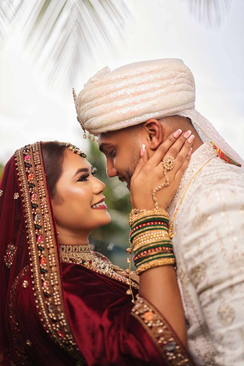 Couple in traditional South Asian wedding attire share an intimate moment with bride in red lehenga and groom in white sherwani.