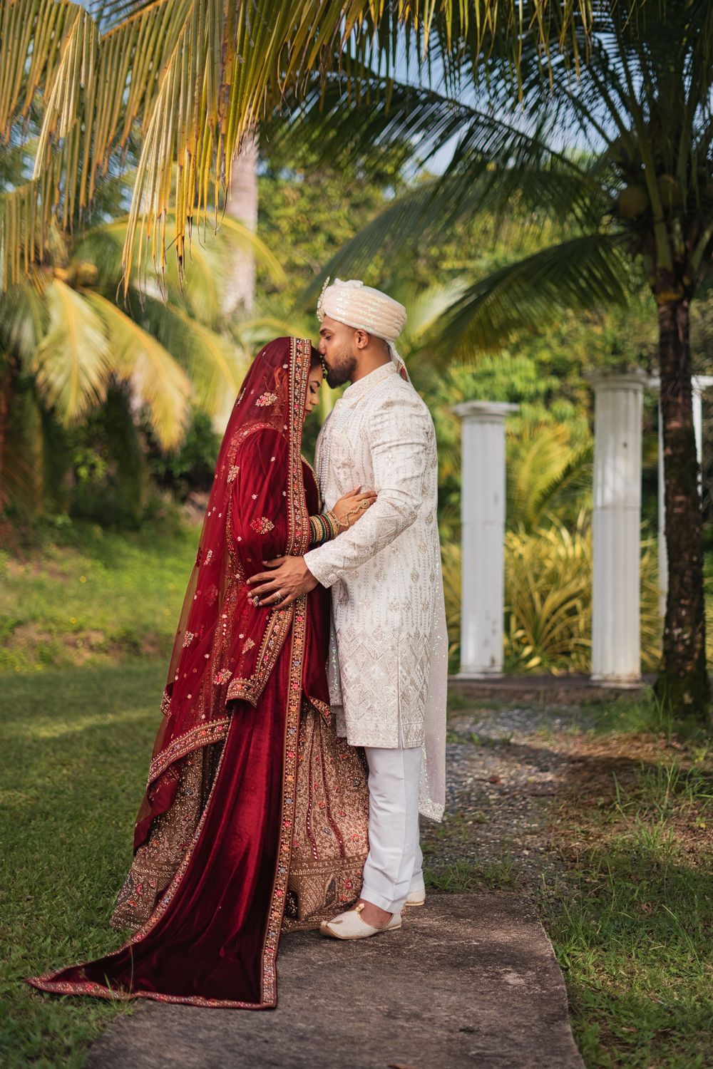A couple in traditional South Asian wedding attire embraces under palm trees in a tropical garden setting.