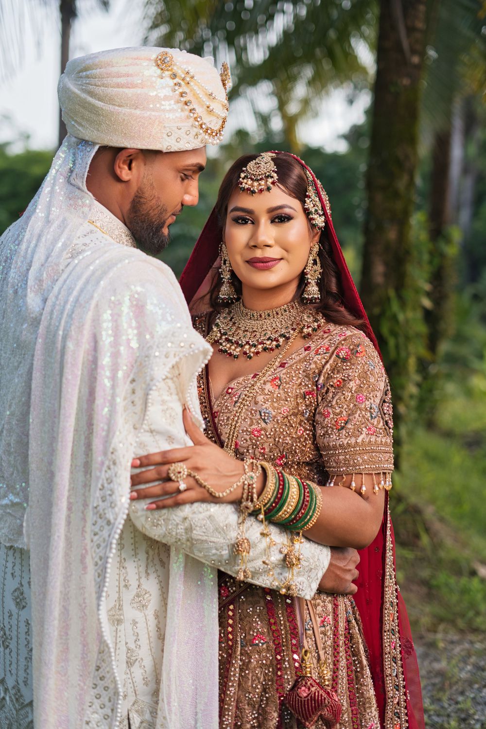 Traditional South Asian couple in ornate wedding attire pose together outdoors with palm trees in background.
