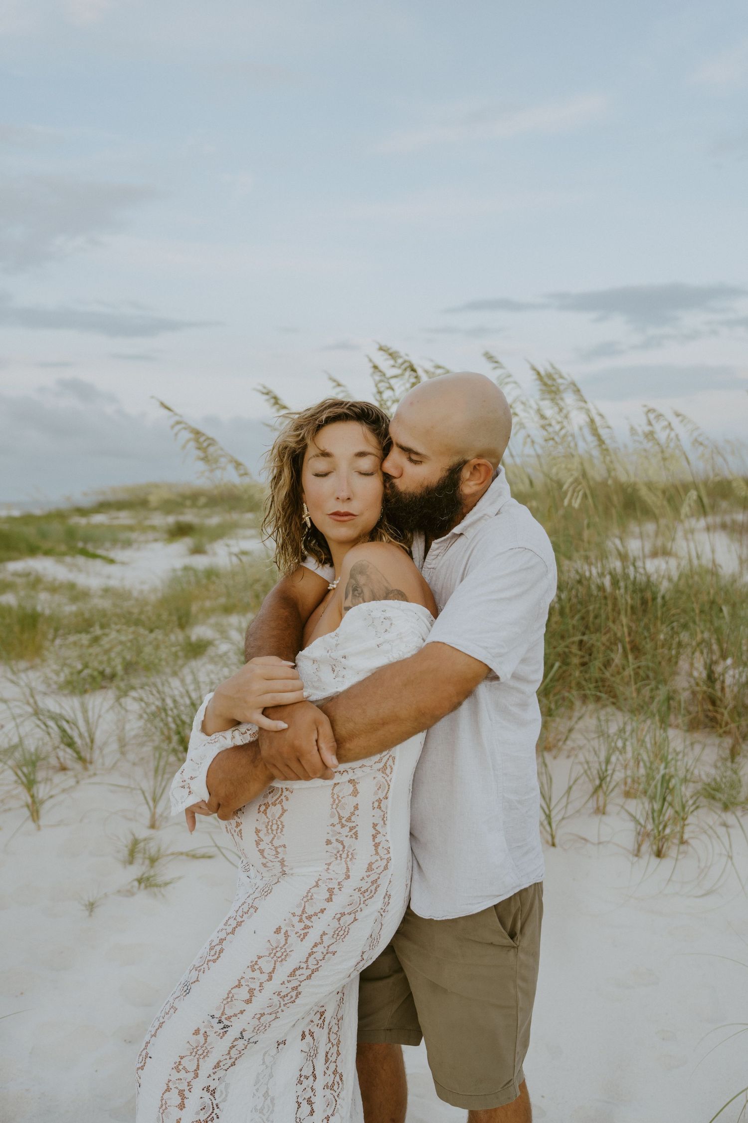 A couple in white attire walks along a sandy beach path at sunset with palm trees in the background.