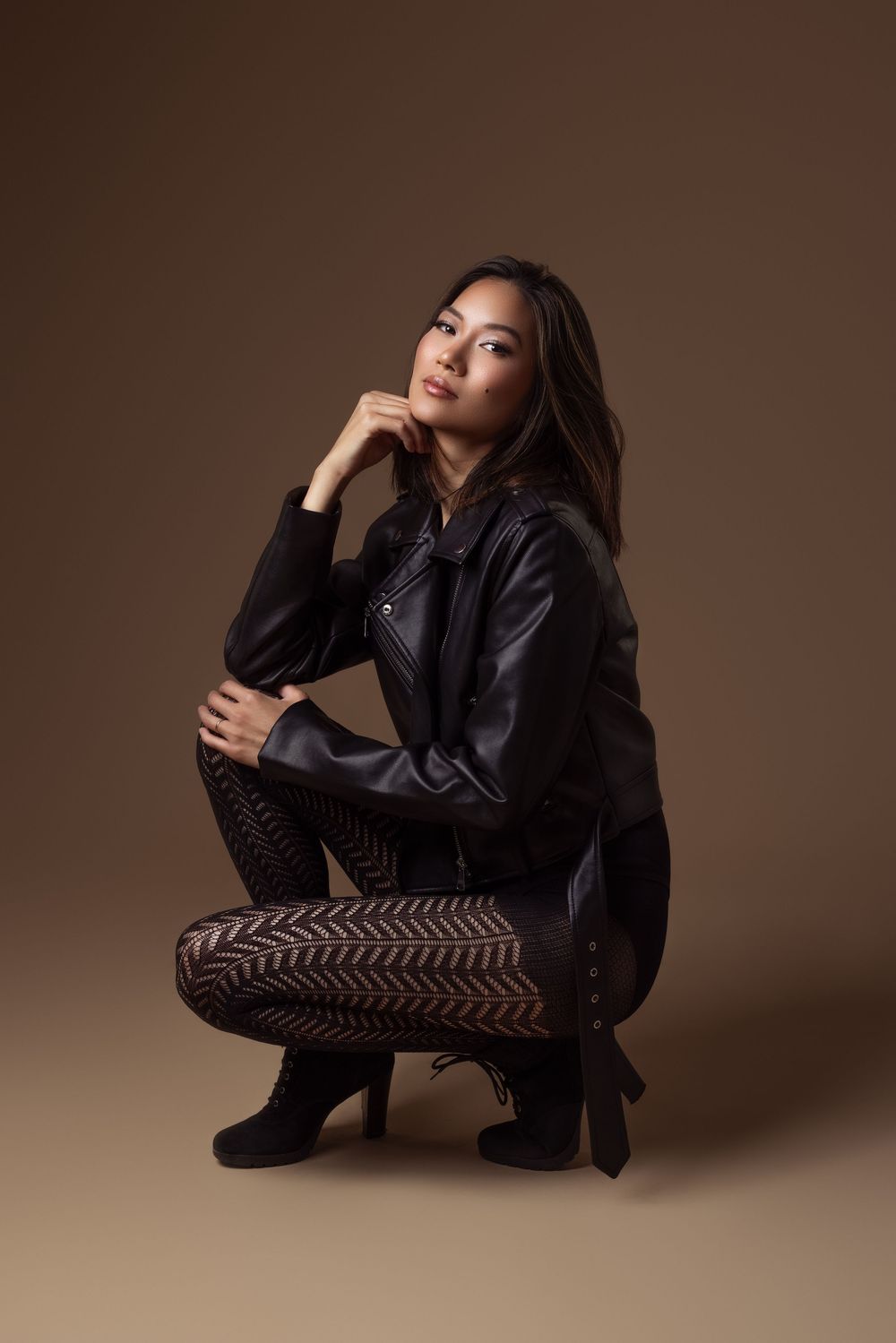 Woman in black leather jacket and patterned leggings crouching gracefully against a warm brown studio background.