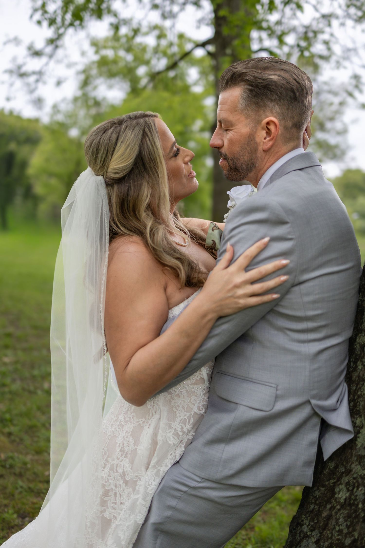 Bride in lace gown and groom in gray suit share a romantic moment leaning against a tree outdoors.