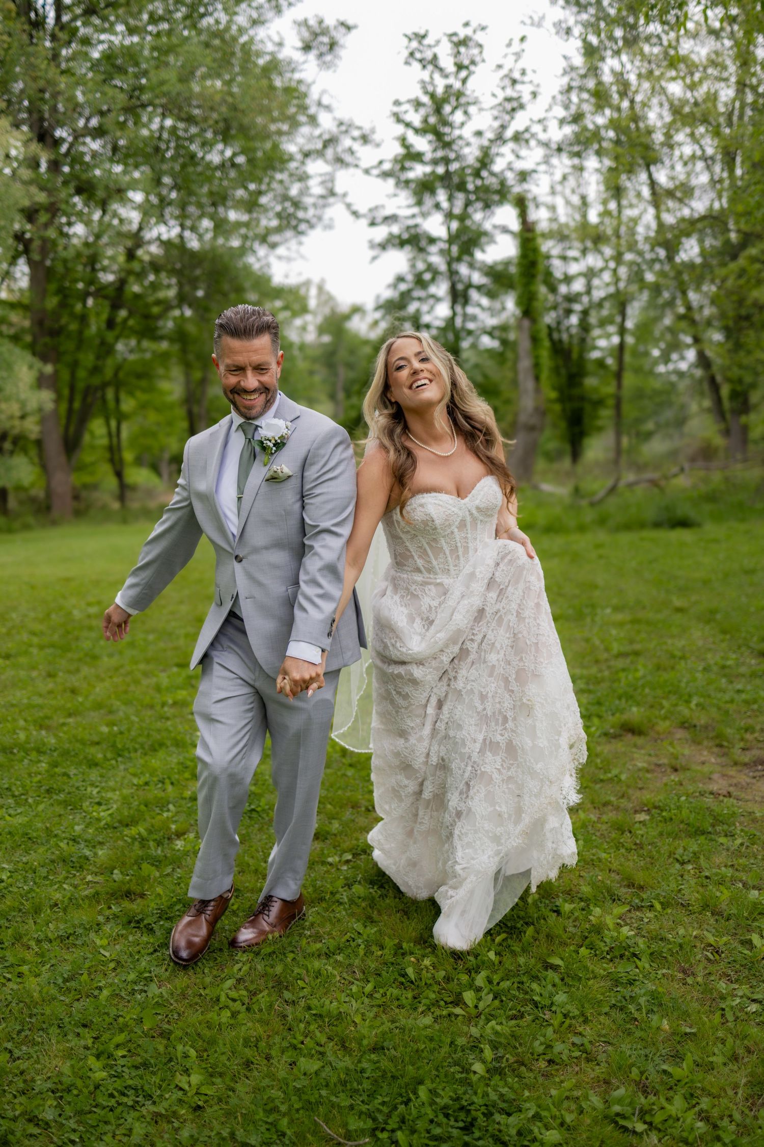 Joyful bride and groom hold hands while walking through a lush green park on their wedding day.