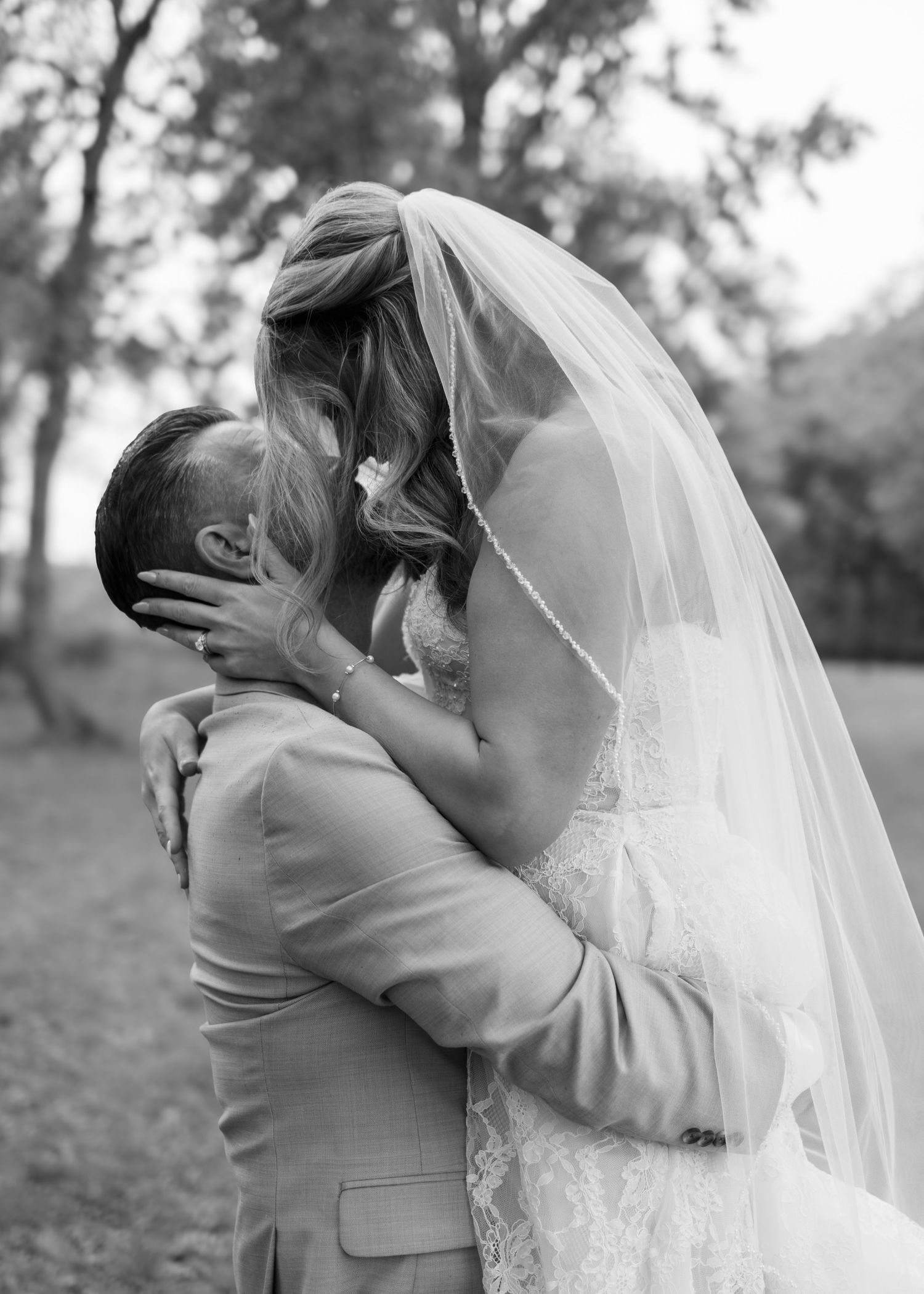 Black and white photo of bride and groom sharing a romantic kiss, bride wearing lace gown and veil outdoors.