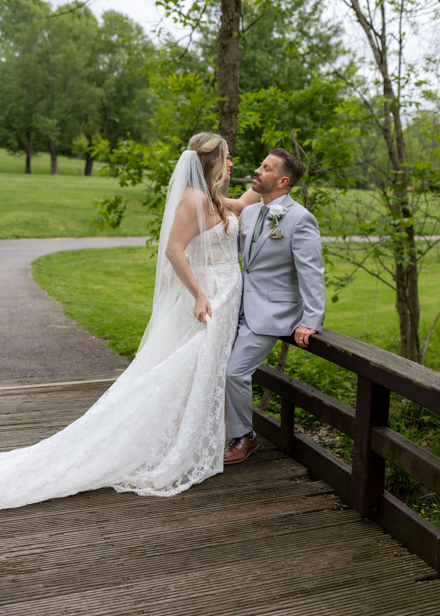 Bride in lace gown and veil with groom in gray suit sharing a romantic moment on a wooden bridge outdoors.