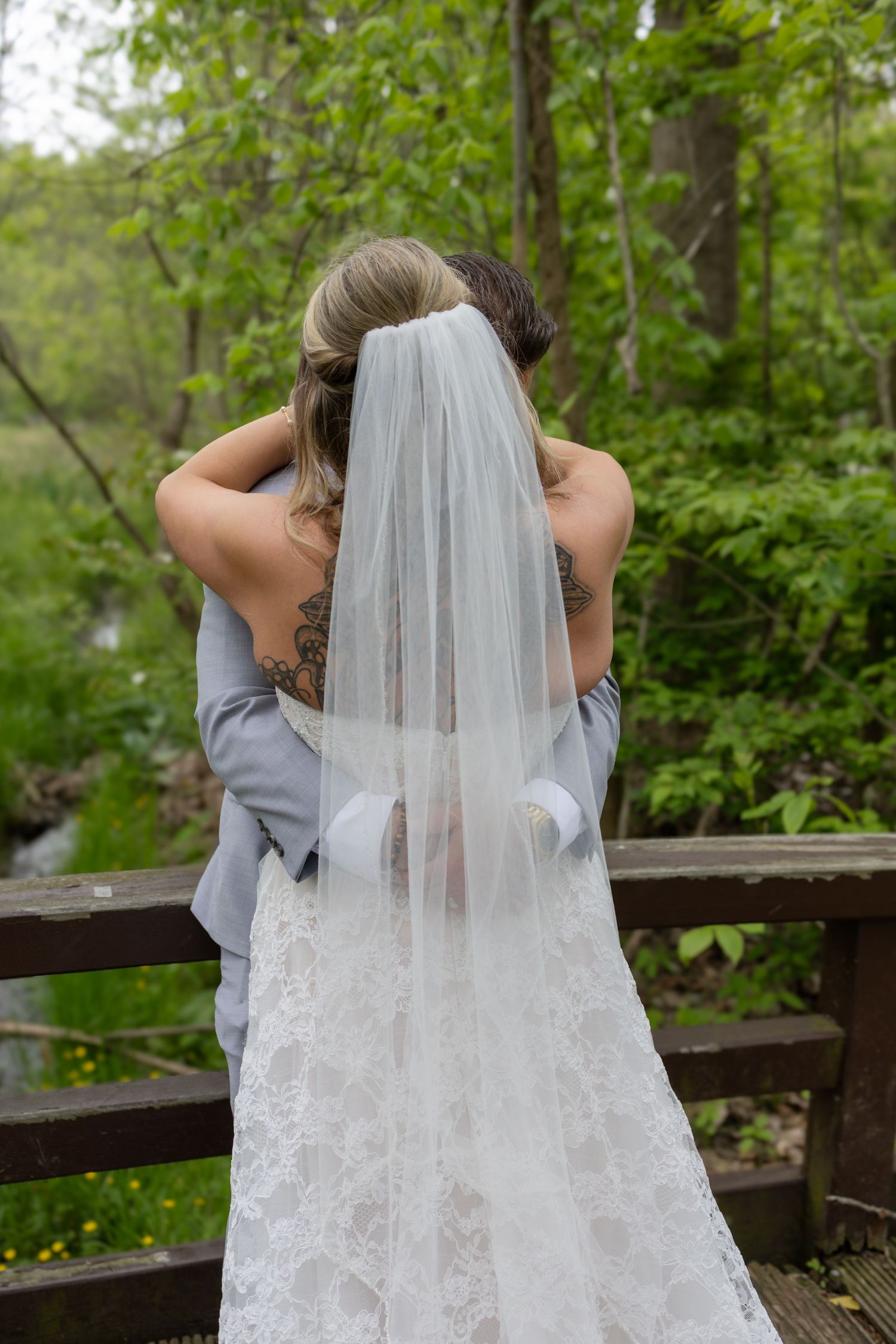 Bride and groom embracing on a wooden bridge, showing the bride's lace dress, veil, and groom's tattoos.