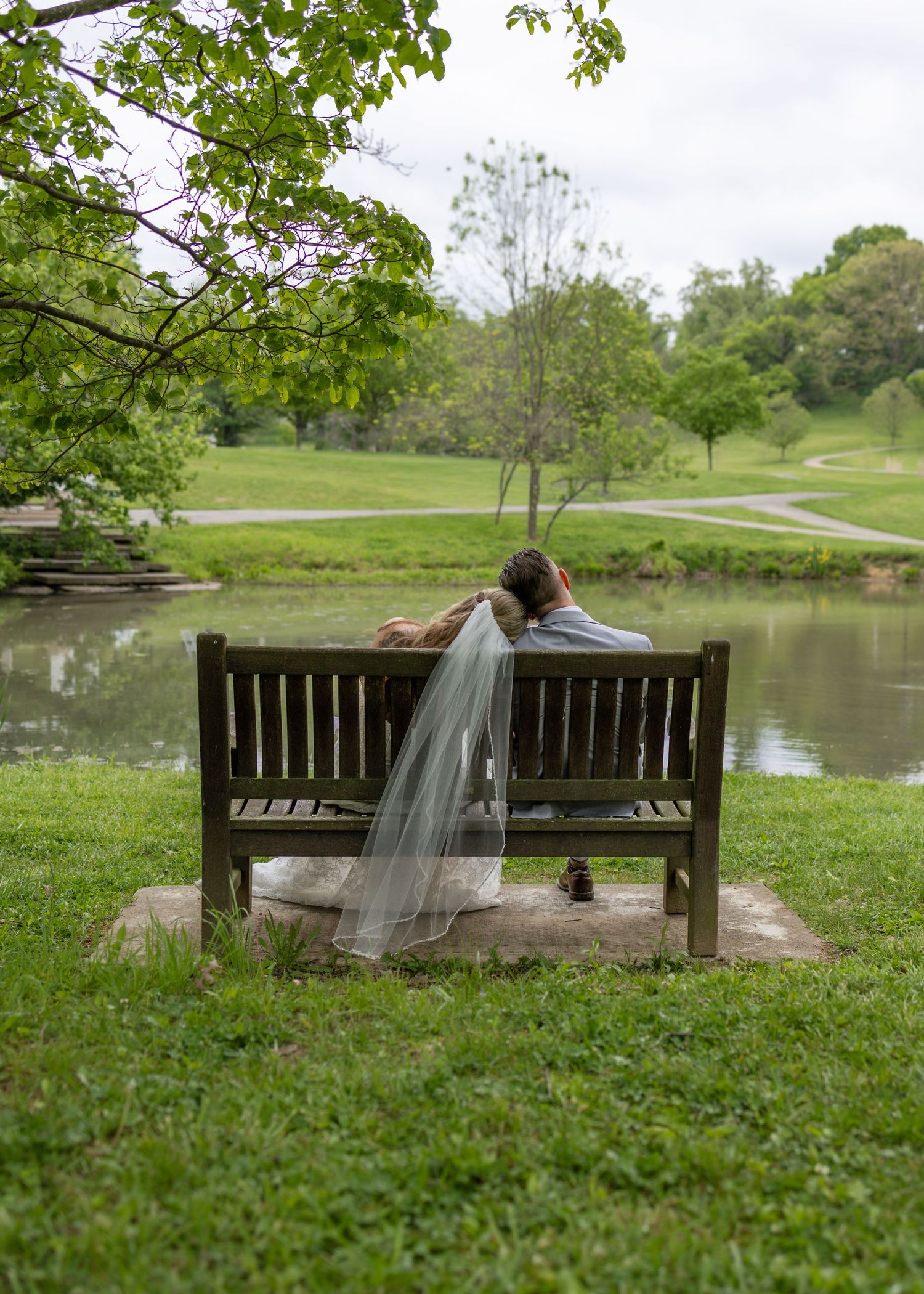 Bride and groom sitting together on a wooden bench overlooking a serene pond in a lush green park.