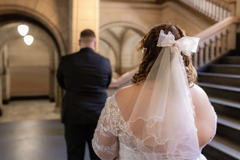 Bride in lace wedding dress and veil with bow stands beside groom in navy suit near grand staircase.