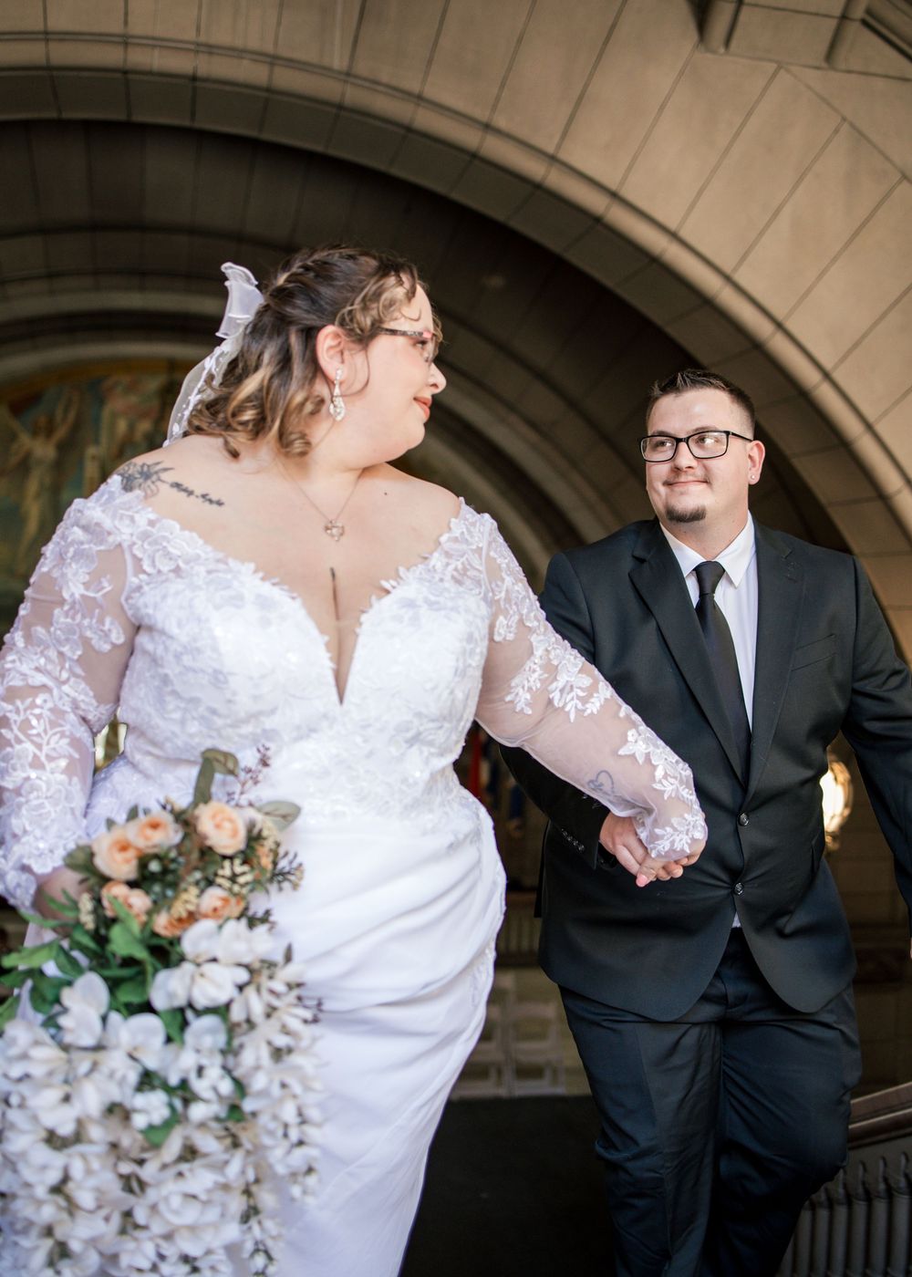 Bride in lace gown holding bouquet walks hand-in-hand with groom in black suit on church steps.