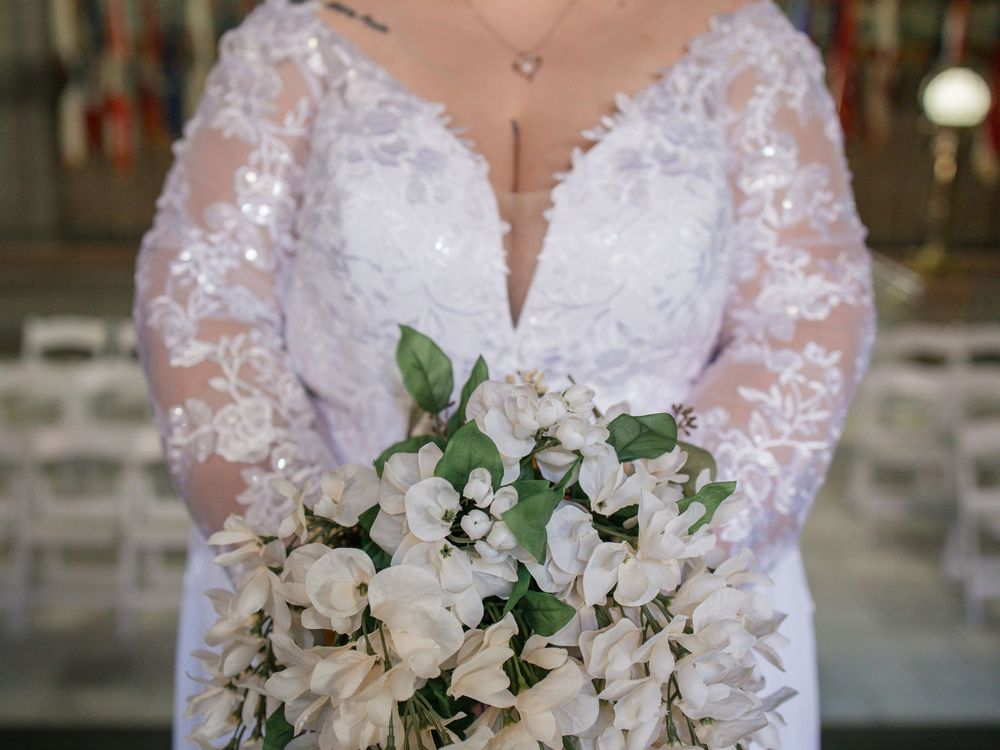 Bride in lace long-sleeve wedding gown holding elegant white floral bouquet with greenery at indoor venue.