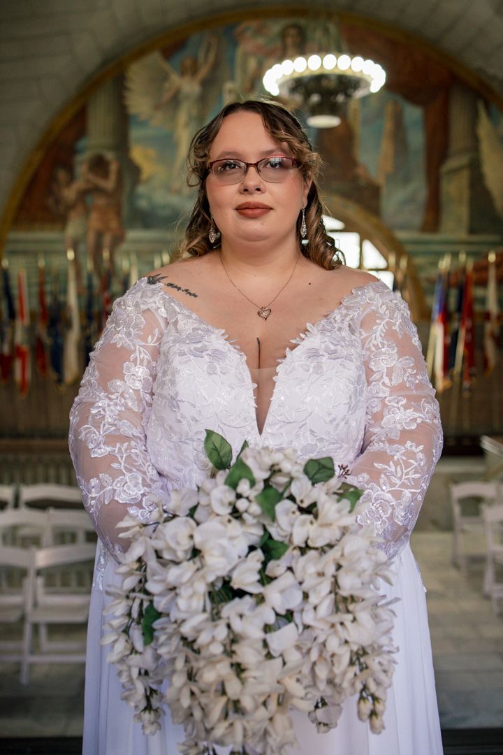 Bride in lace long-sleeve wedding gown holds white cascading bouquet inside ornate ceremony hall with murals.