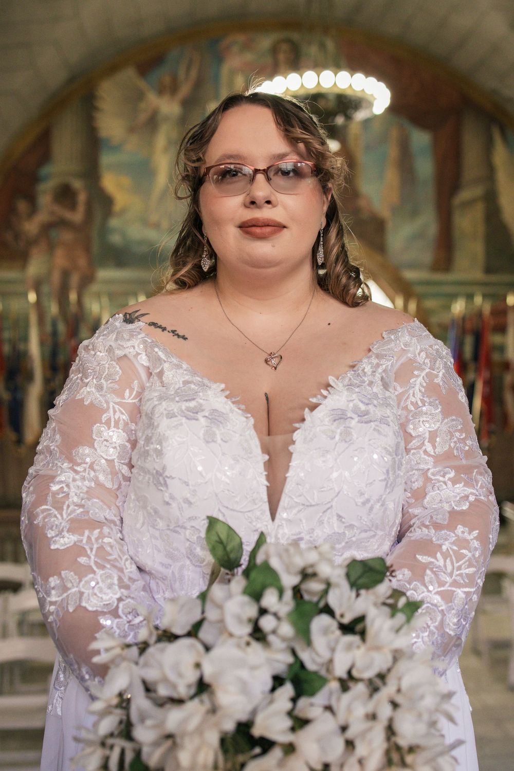 Bride in lace long-sleeve wedding gown holding white bouquet, standing in ornate church with mosaic backdrop.
