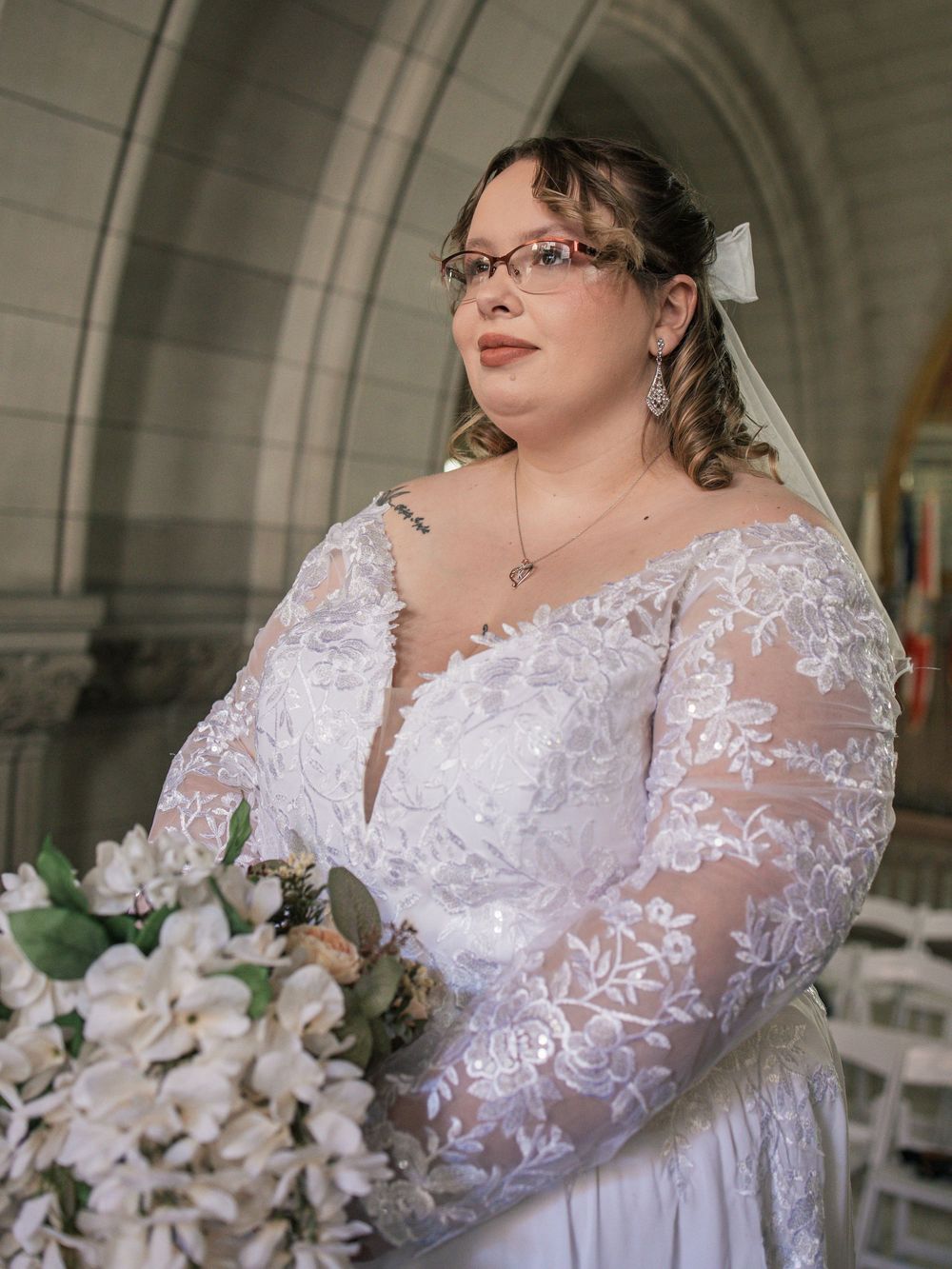 Plus-size bride in lace long-sleeve wedding gown holding white floral bouquet, wearing glasses and veil in church.