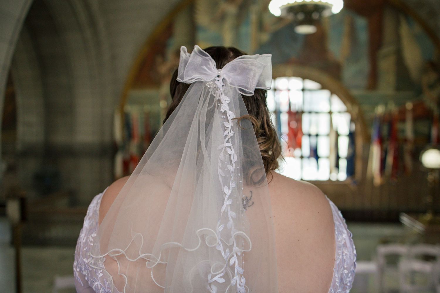 Bride wearing a white wedding veil with bow detail, standing in a church with stained glass windows.