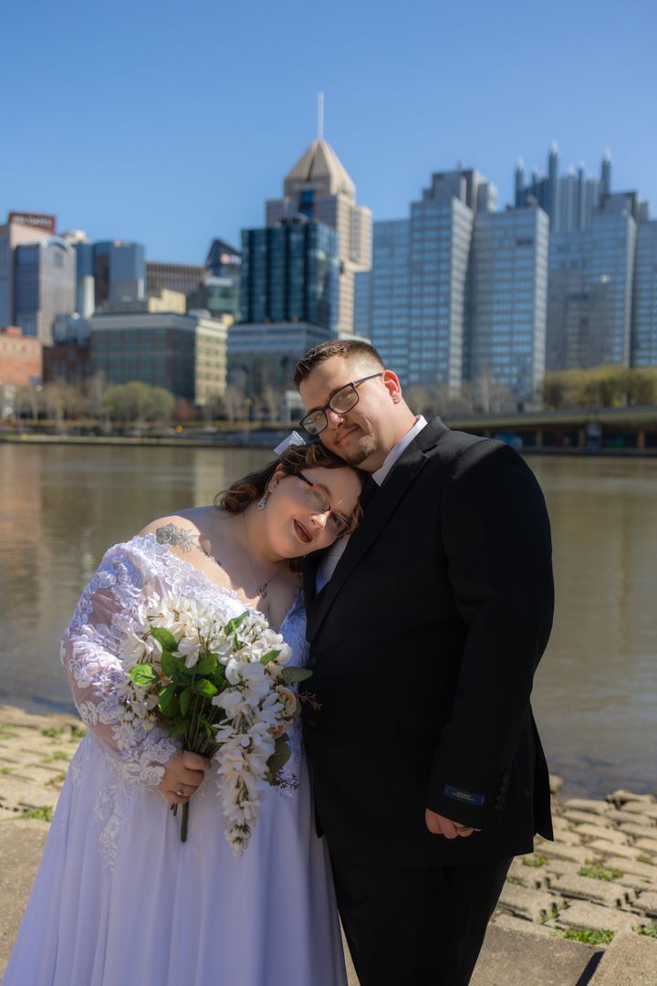 Bride and groom pose romantically by a river with a city skyline backdrop on a sunny day, bride holding white bouquet.