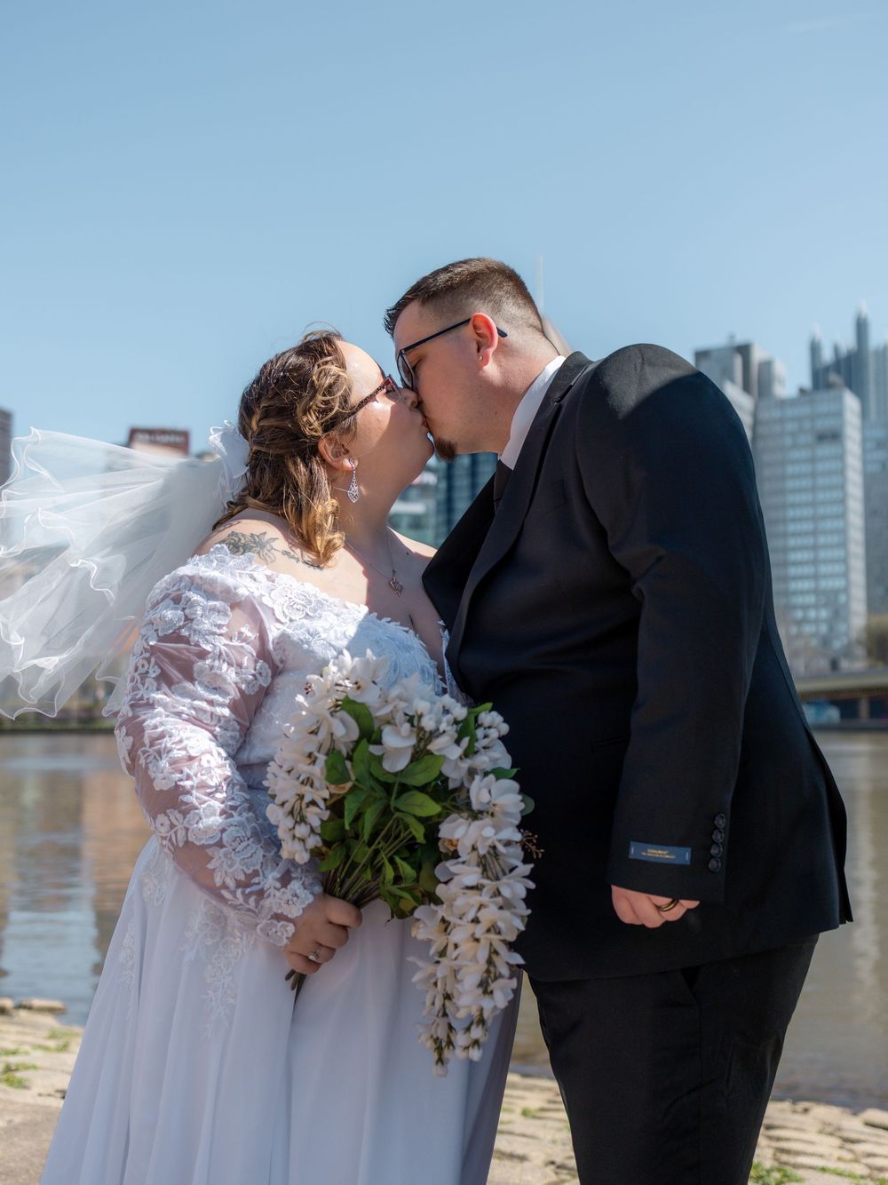 Newlywed couple sharing a kiss in wedding attire by a riverside with a city skyline on a sunny day.