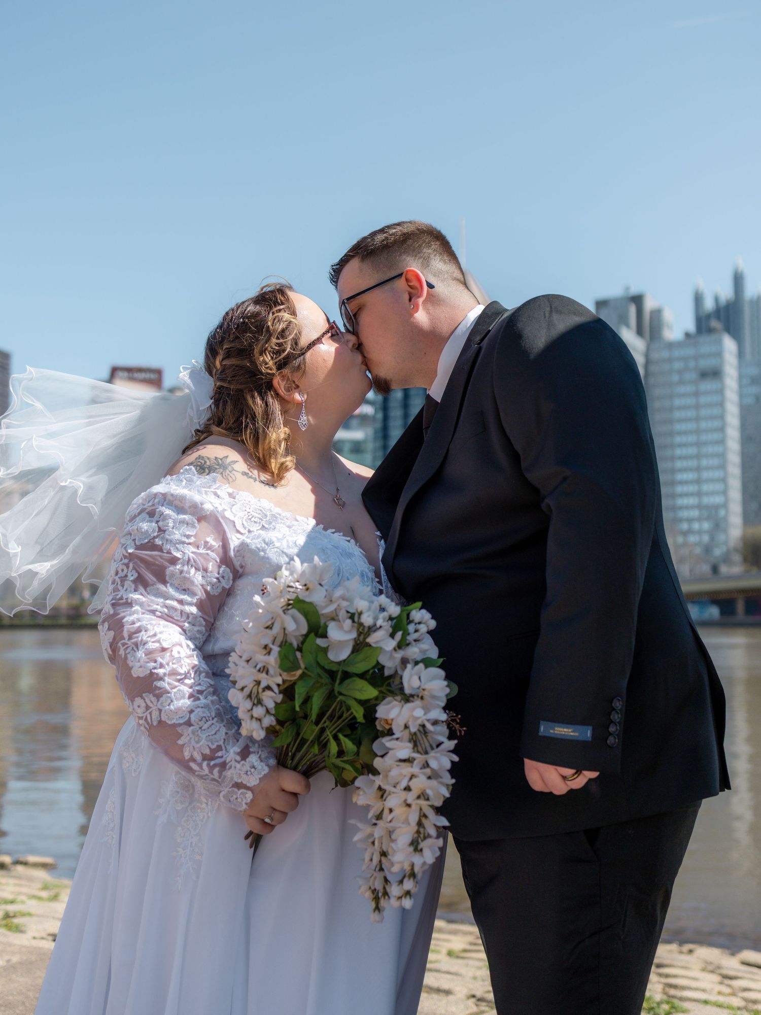 Newlywed couple sharing a kiss in wedding attire by a riverside with a city skyline on a sunny day.